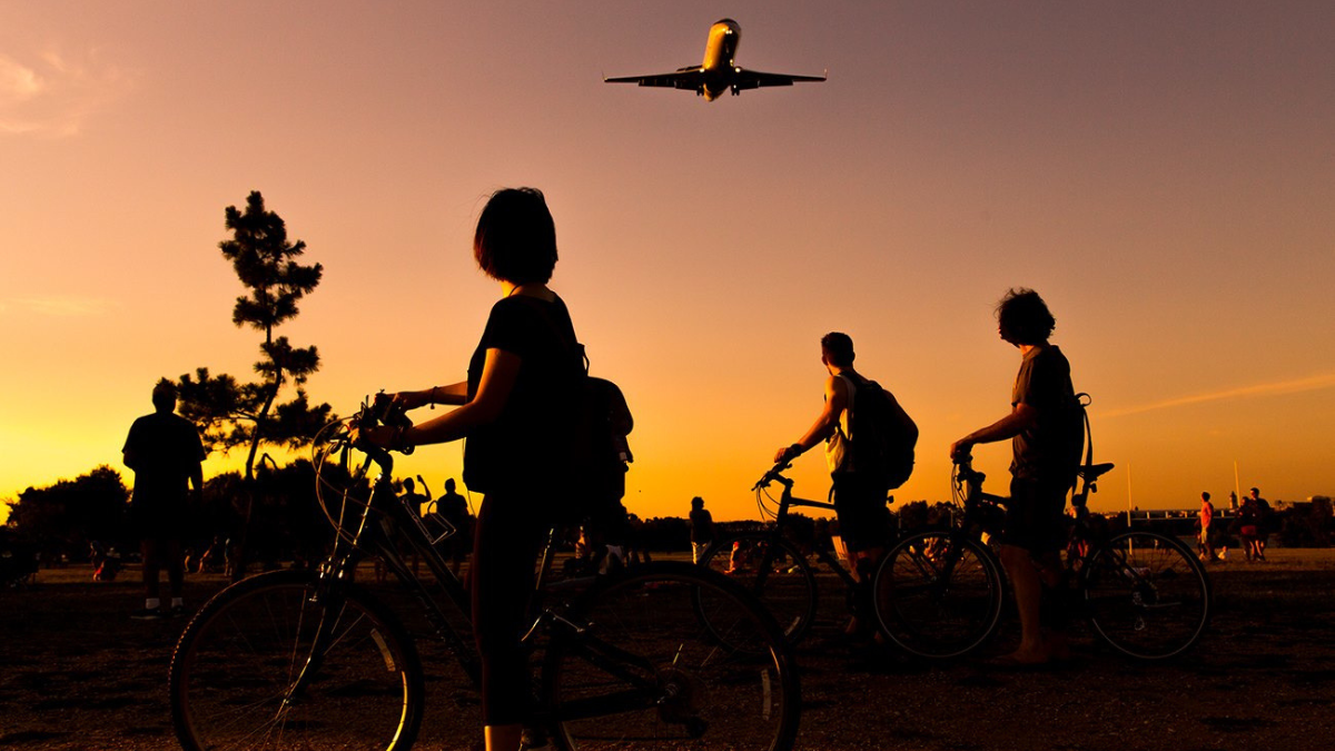 People riding bikes at a park stop to watch a plane fly overhead 