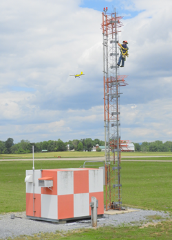 Technician climbing glide scope on airfield with general aviation aircraft in the background.