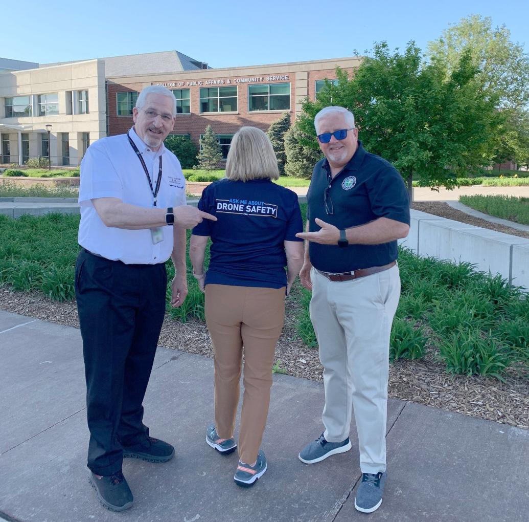 "image showing two men and a woman with her back turned showing the back of her shirt saying "drone safety" on the logo"