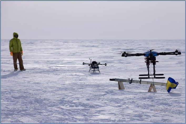 man standing on left, two drones taking off on snow coevered ground